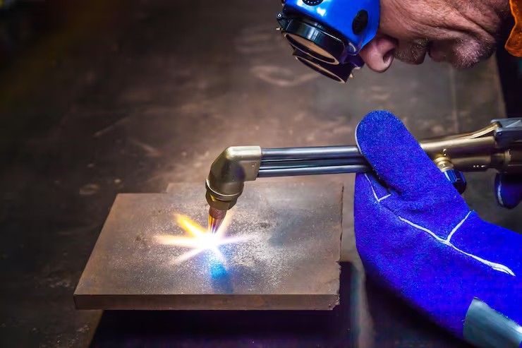 Metalworker with blue safety gloves using an oxy-fuel cutting torch on a thick metal plate, bright flame